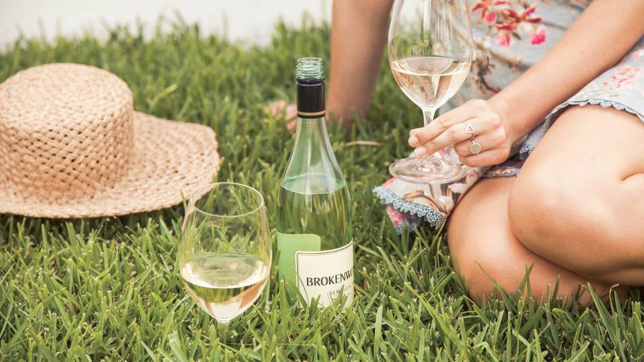 Woman sitting on a grass lawn serving herself a white wine from Brokenwood