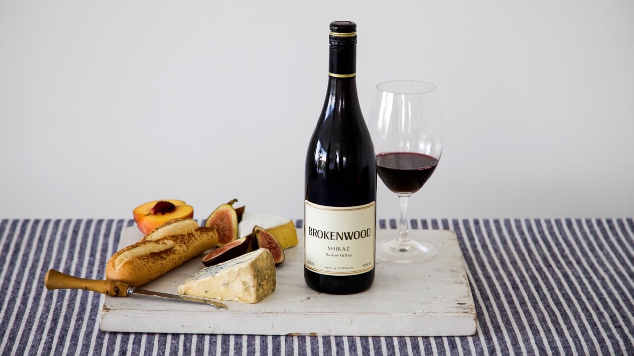 A bottle and glass of Brokenwood Shiraz with a cheese board on a striped tablecloth