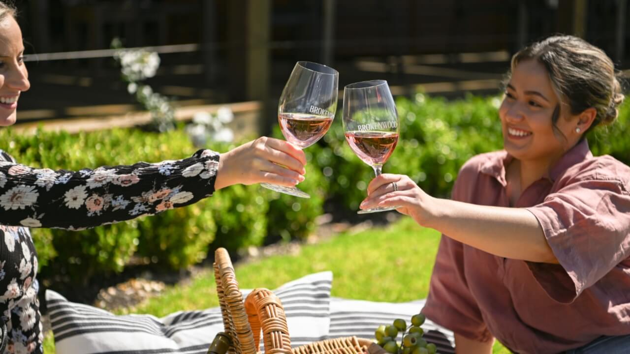 Two women toasting glasses of wine on the lawn of Brokenwood’s Cellar Door